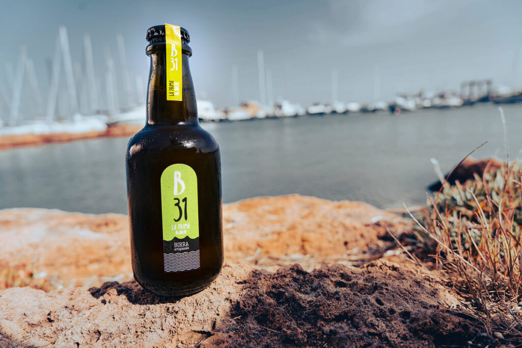 Bottiglia di Blanche B31 in primo piano, con paesaggio marino e barche sullo sfondo.